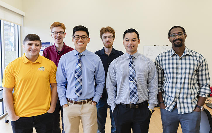 Six male students standing in front of the camera smiling in a well lit room 