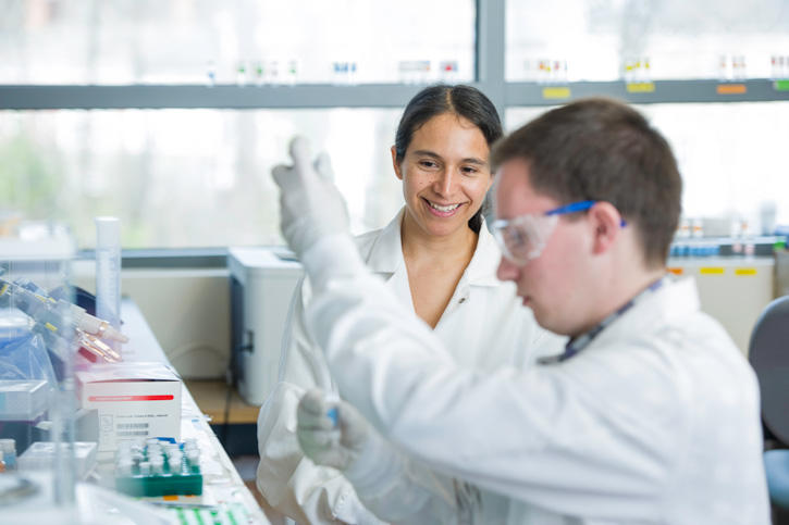 Two people in lab coats, one with lab goggles on, pipetting test materials.